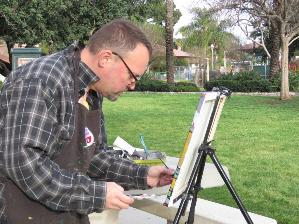 Artist Oscar Will painting on a canvas on an easel, paintbrush in left hand, hunched over slightly and concentrating really hard on what he's doing. He is wearing a black smock and a grey checkered jacket. In his right hand is a small plastic palette with paint on it. His painting supplies sit to his left on a picnic table and in the background grass and trees from the park-like setting.
