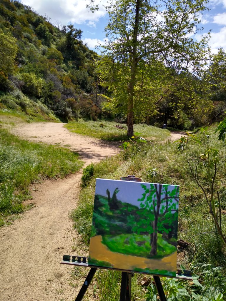 Painting on an easel next to a dirt path that forks around a grass field with a tree in it.